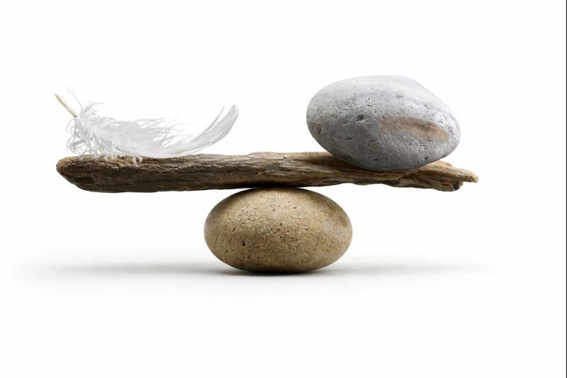 Una pietra liscia che sostiene un legnetto orizzontale. Una piuma bianca (leggerezza) a sinistra e un sasso grigio (pesantezza) a destra.